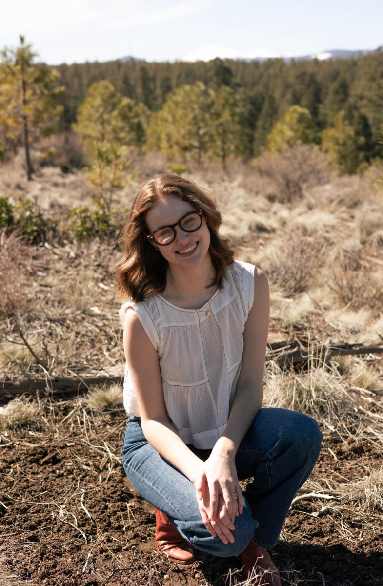 Portrait of a smiling white woman with brown hair wearing glasses, crouched in front of grasslands with trees in the background.