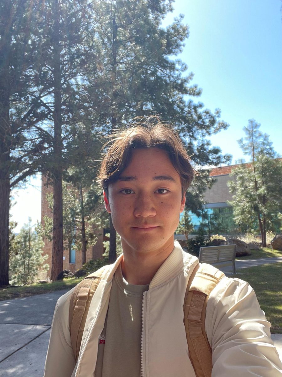 Selfie of a young Asian American man wearing a white jacket and tan backpack in front of pine trees and a modern building.